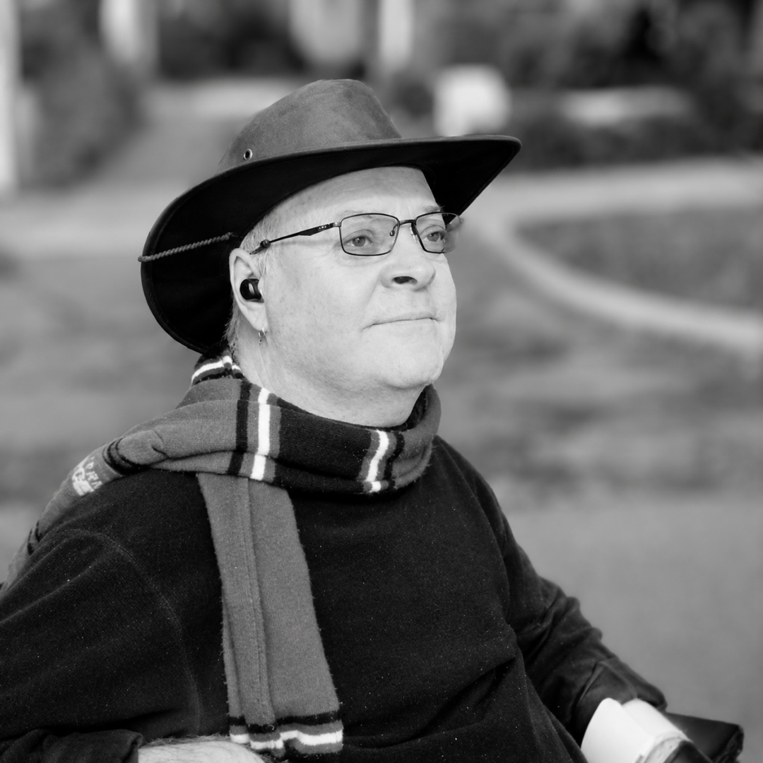 Darren West, a man in a wide-brimmed hat, glasses and a striped scarf, seated outdoors.